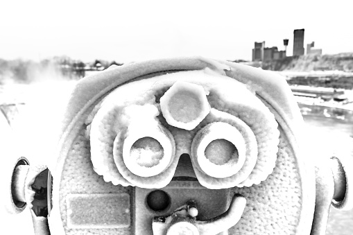 Monochrome image of an ice-covered viewfinder at Niagara Falls, NY.  To the right-hand, in the background, is Niagara Falls, Ontario (Canada) and to the left-hand in the background are the American Falls in Niagara Falls, NY.  A tower viewer is a set of binoculars permanently mounted on a pole and are usually coin operated.