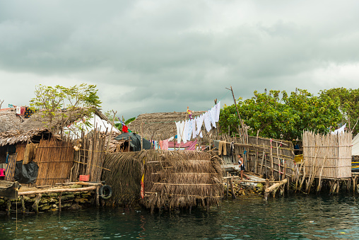 Some simple homes made of wood and bamboo sticks on the water's edge against cloudy sky. A woman hangs laundry. View from the sea. San Blas village of Kuna Yala ethnic group. Panama