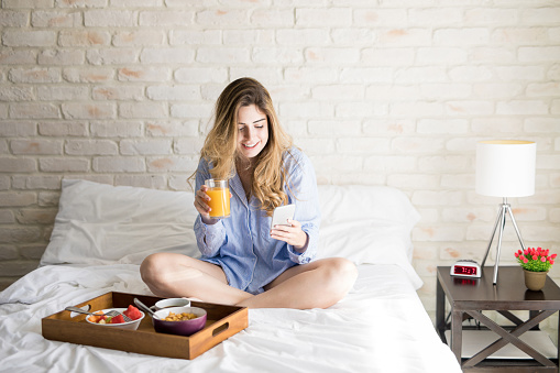 Cute Hispanic woman sending a text on her smartphone while eating breakfast in bed