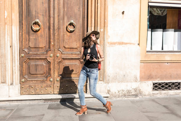 Side view of beautiful young woman walking on streets and taking pictures. Wears casual clothes, sunglasses, a hat and a backpack.