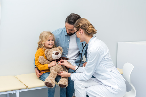 female doctor listening to breath of teddy bear of little girl