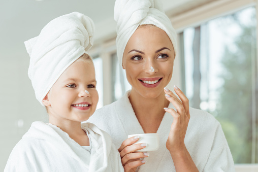 beautiful happy mother and daughter in bathrobes and towels applying face cream together