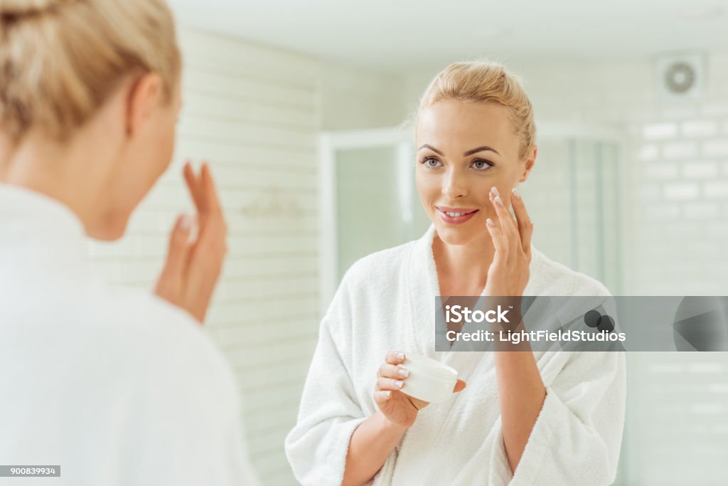 woman in bathrobe applying face cream beautiful smiling young woman in bathrobe applying face cream Face Cream Stock Photo