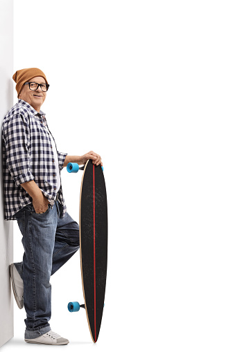 Full length portrait of an old hipster with a longboard leaning against a wall isolated on white background