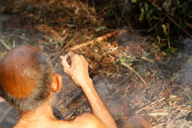 an elderly Thai Theravada Buddhist Monk sitting by a recently lit, smoking fire in the temple grounds in Thailand, Southeast Asia