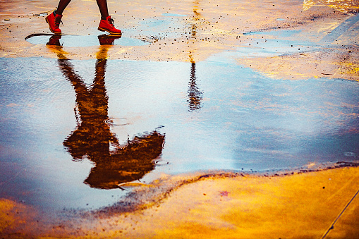 Reflection in a puddle of a young woman holding umbrella, walking outdoors over an empty yellow parking lot on a cold, windy, rainy autumn day.
