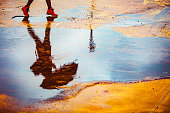 Reflection in a puddle of a young woman holding umbrella, walking outdoors on a cold, windy, rainy autumn day.