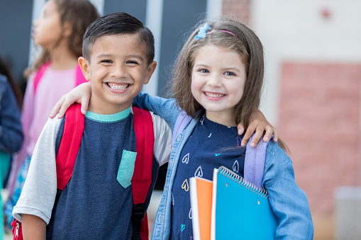 Cute adorable schoolboy and schoolgirl smile as they  stand outside the school building. They are waiting for school to start. Their arms are around each other.