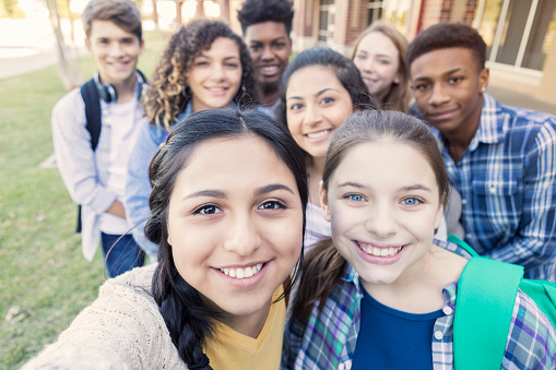 Diverse group of eight high school students are smiling and looking at the camera. Teenagers are students at public high school, and are wearing backpacks or holding school books.