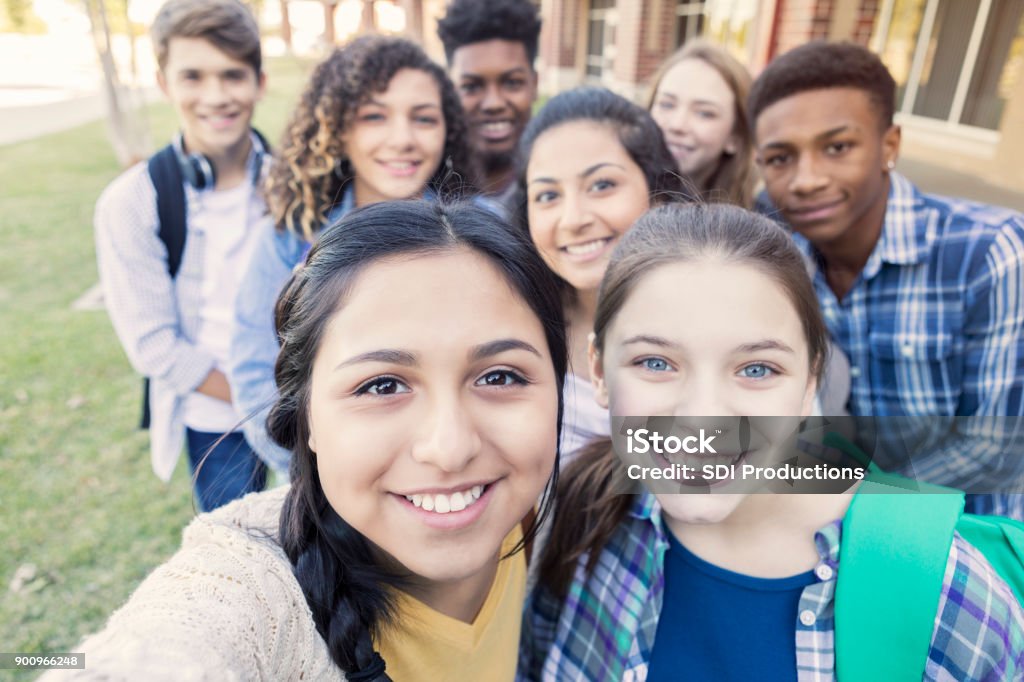 Diverse group of teens looking at camera taking selfie at high school Diverse group of eight high school students are smiling and looking at the camera. Teenagers are students at public high school, and are wearing backpacks or holding school books. Teenagers Only Stock Photo