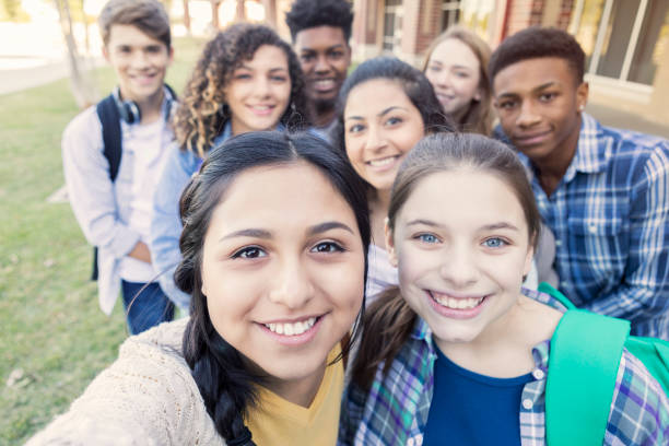 Diverse group of teens looking at camera taking selfie at high school Diverse group of eight high school students are smiling and looking at the camera. Teenagers are students at public high school, and are wearing backpacks or holding school books. teenagers only photos stock pictures, royalty-free photos & images