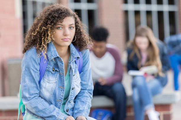 Sad teen girl looking at camera outside high school Pretty biracial teenage girl is smiling and looking at the camera. She is sad or depressed and is sitting outside a public high school. Student is wearing a backpack and classmates are sitting behind her. boy and girl looking at camera stock pictures, royalty-free photos & images