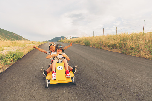 Two young boys wearing racing caps and flight goggles race a homemade car down the road. The driver concentrates on driving while the other boy raises his arm in successful triumph. They have found speed. Image taken in Utah, USA.