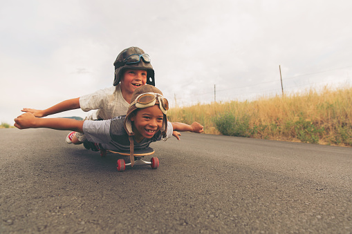 Two young boys dressed in flight caps and goggles ride down a hill on their skateboard hoping to take flight. Their imaginations of being pilots and piloting an aircraft are not too far off. Image taken in Utah, USA.