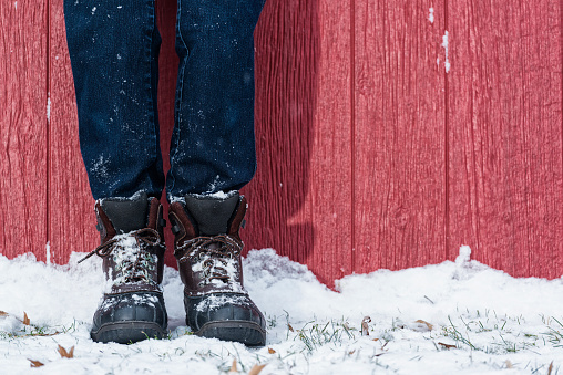 A close-up of a woman's legs wearing duck boots and blue jeans leaning against a red barn in the snow.