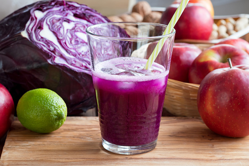 Purple cabbage juice in a glass, with cabbage, apples and a lime in the background