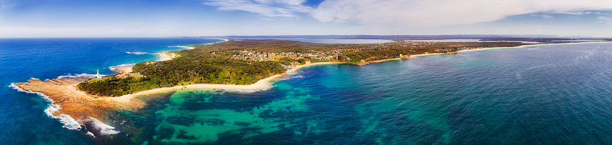 Norah head lighthouse and nature reserve on Central coast of NSW in wide aerial panorama. Clear pacific ocean water breaks at sandy beaches and seafloor of continent shore.