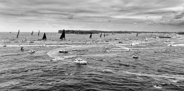 Super maxi leading yachts clearing from Sydney harbour at start of Sydney-Hobart yacht regatta surrounded by crowd of boats and ferries in wide BW aerial.