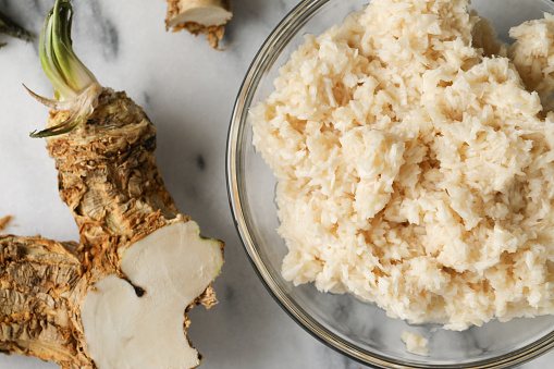 An overhead, extreme close up horizontal photograph of some homegrown horseradish roots and a glass bowl of prepared horseradish on a marble countertop.