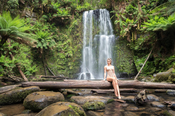 hermosa mujer sentada en un tronco en frente de una impresionante cascada, naturaleza, cataratas de beauchamp, great ocean road - rainforest waterfall australia forest fotografías e imágenes de stock