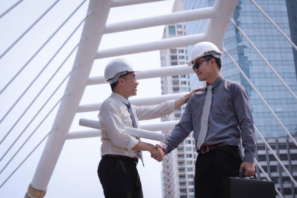istock Asian businessman discuss and shake hand with engineer architect professional wearing safety helmet and suite, holding construction roll plan and briefcase for working team concept 901060110