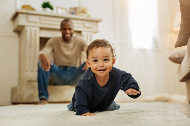 Smiling father watching his son crawling Happy childhood. Happy dark-haired afro-american man laughing and watching his cheerful young son crawling on the floor crawling photos stock pictures, royalty-free photos & images