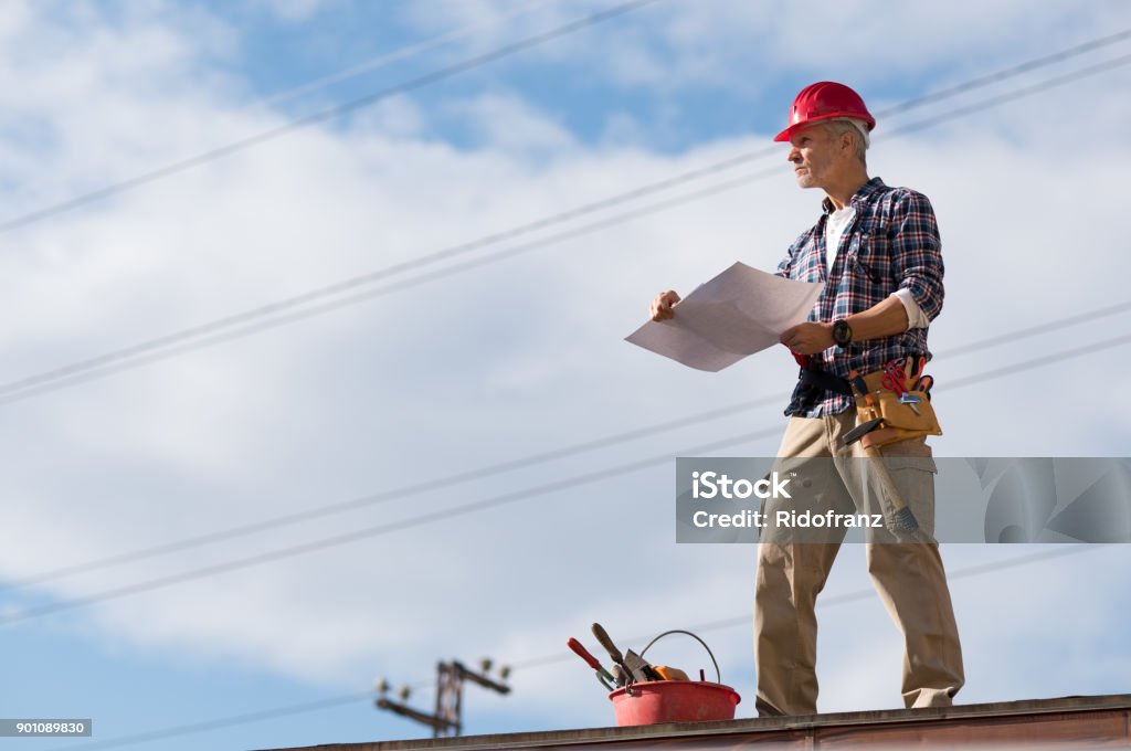 Construction worker on rooftop Engineer standing on rooftop looking away and holding blueprint project. Mature construction worker kit standing on rooftop with copy space. Workman inspecting construction site with sheet of paper in hand. Rooftop Stock Photo