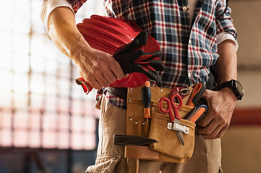 Closeup of bricklayer hands holding hardhat and construction equipment. Detail of mason man hands holding work gloves and wearing tool kit on waist. Handyman with tools belt and artisan equipment.