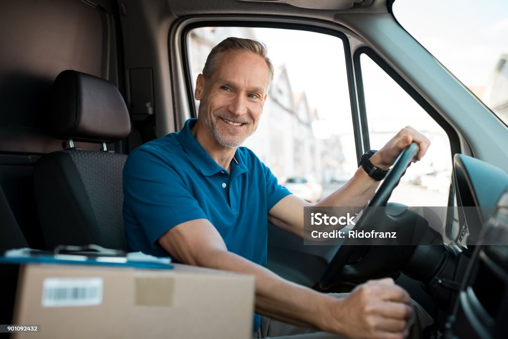 Delivery man driving van Delivery man driving van with packages on the front seat. Happy mature courier in truck. Portrait of confident express courier driving his delivery van. Van - Vehicle Stock Photo