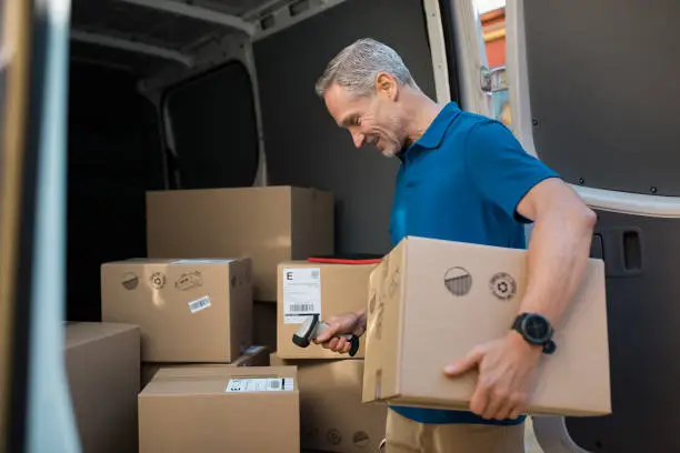 Delivery man scanning cardboard boxes with barcode scanner. Courier holding parcel and scanning barcode with barcode reader in van. Mature man reading and scanning labels on boxes before shipment.
