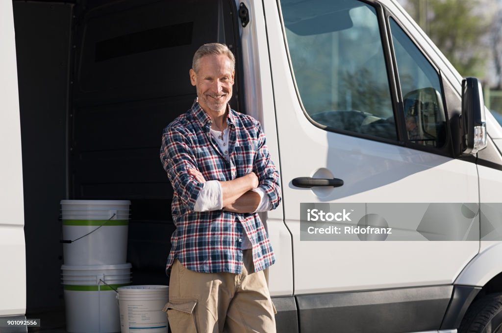 Painter standing against van Portrait of painter standing next to his van with paint container. Decorator standing next to van and looking at camera. Happy smiling worker leaning on his white truck with folded arms. Craftsperson Stock Photo