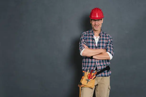 Successful bricklayer with red helmet and equipment tool kit on waist standing against grey wall. Portrait of happy manual worker isolated over grey background. Satisfied mature craftsman with copy space.