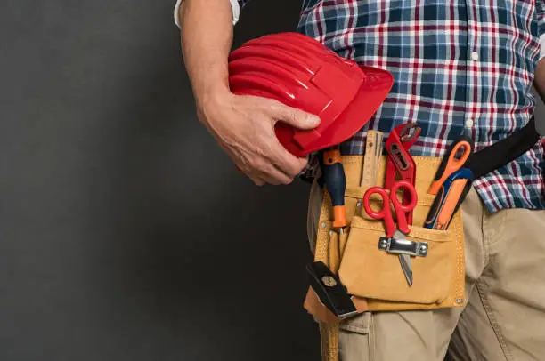 Closeup of hardhat held by construction worker on grey background. Detail of bricklayer holding red helmet and kit tool. Closeup of craftsman hand holding tool belt with equipment on grey wall with copy space.