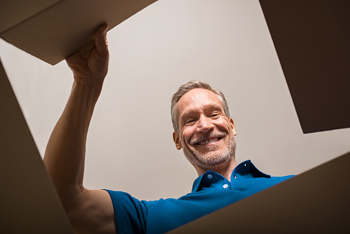 Happy mature man looking into parcel cardboard box and smiling. Cheerful senior man happy on seeing package. Smiling man feeling overjoyed on seeing parcel and opening it.