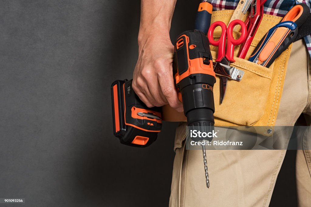 Hand holding construction tools Close up of handyman holding a drill machine with tool belt around waist. Detail of artisan hand holding electric drill with tools isolated over grey background. Closeup hand of bricklayer holding carpentry accessories. Craftsperson Stock Photo