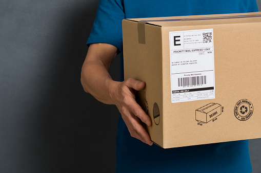 Closeup hands of delivery man holding package to deliver. Courier hand holding brown box isolated on grey background. Detail of delivery man carrying cardboard parcel with label with copy space.