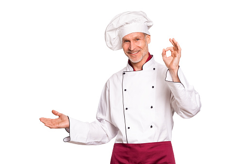 Smiling senior cook showing something isolated on white background. Portrait of happy mature chef presenting over white background. Cheerful chef man looking at camera isolated on white with hand sign ok.