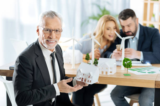 istock Smiling man showing a miniature house while his colleagues working at windmills 901097748