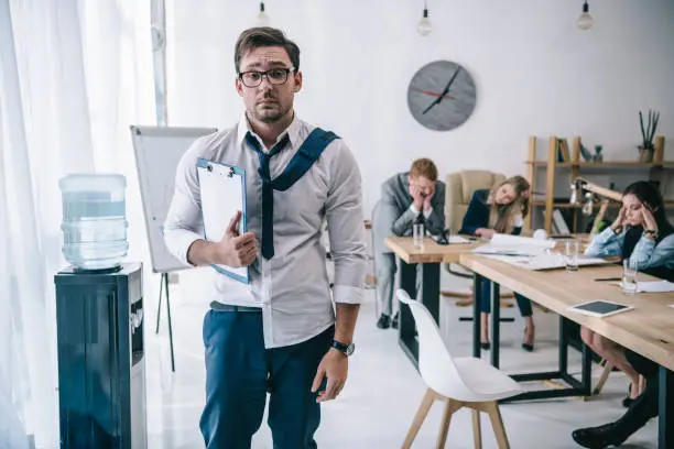 overworked untidy businessman with clipboard standing at office while colleagues sitting at conference hall