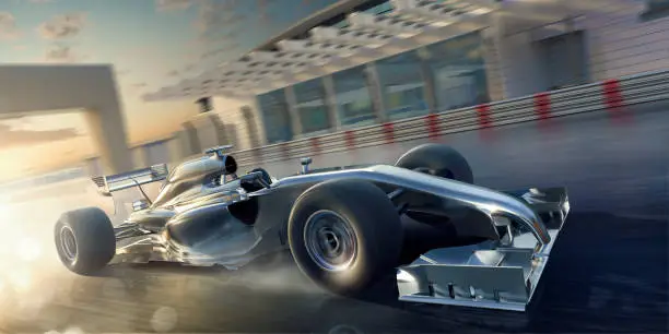 A close up image of a generic silver unmarked racing car, moving at high speed in slightly wet conditions. The racecar is being driven along a straight section of racetrack on a generic racetrack, past outbuildings and safety barriers at dusk.
