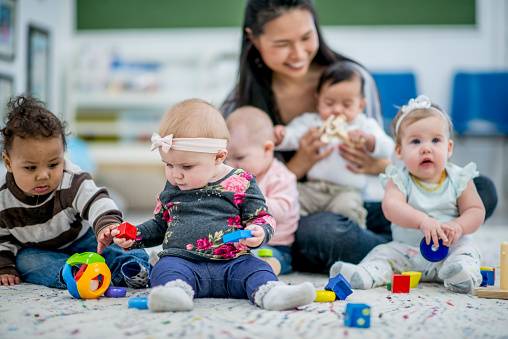 A group of babies are indoors in a daycare center. They are sitting on the carpet and playing with colorful wooden blocks and a ball. The babysitter is watching over them.