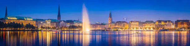 Sweeping panoramic vista across the blue waters of Binnen-Alster lake and its famous fountain to the warm lights of the stores, hotels and restaurants on Jungfernstieg in the heart of Hamburg, Germany’s vibrant second city.