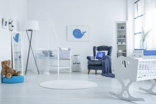 Teddy bear on blue pouf and white cradle in bright kid's bedroom with lamp, carpet and armchair