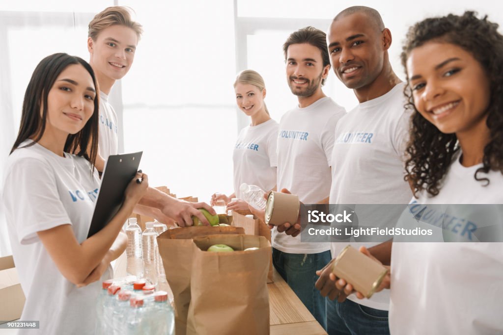 multiethnic group of volunteers multiethnic group of volunteers putting food and drinks in paper bags and looking at camera Volunteer Stock Photo