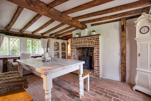 Tastefully modernised dining room withing 16th Century English cottage with traditionally styled decor sympathetic to the original period with dining table and bench stool, cast iron stove, upright clock, exposed timber and beams, brick floor and leaded windows