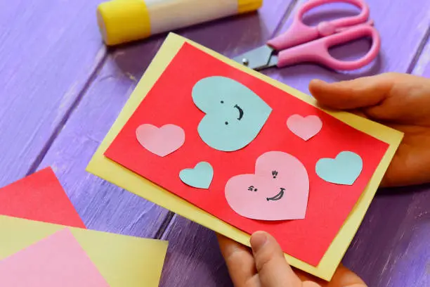 Photo of Child is holding a Valentines card in his hands. Child is showing a greeting card. Happy Valentines Day card. Easy paper crafts for kids concept. Closeup