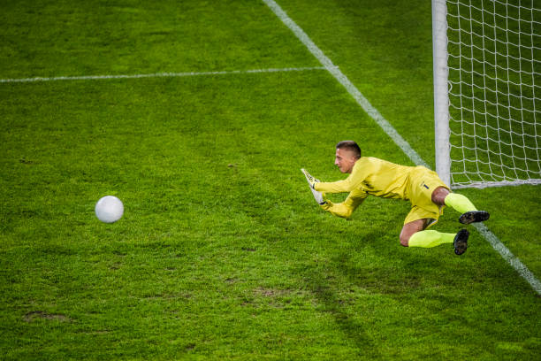 Side view of a soccer goalie in mid-air throwing himself in front of a ball to defend the goal.