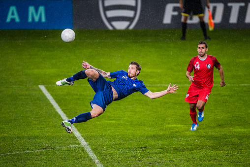 Soccer player in a blue jersey in mid-air in the penalty area about to perform a volley shot.