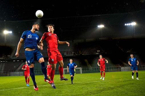 Soccer player in blue jersey in the air heading the ball. Stadium in the background.