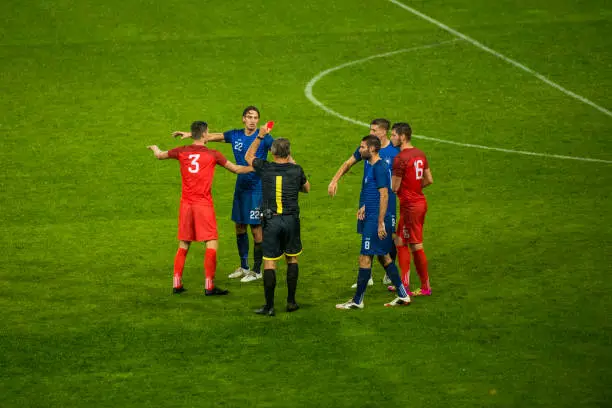 Soccer referee surrounded by players of both teams while he is holding a red card.